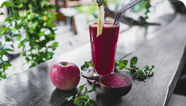 Ein Glas roter Saft mit einem Strohhalm, einem Apfel und einer halben Rübe auf einem Holztisch, umgeben von Pflanzen.