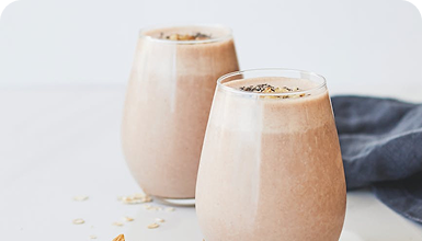 Zwei Gläser mit schokoladigem Smoothie, dekoriert mit Haferflocken, auf einem hellen Tisch.